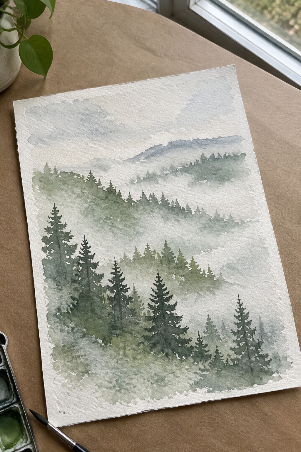 Watercolor misty pine forest mountains painting on table with palette and brush