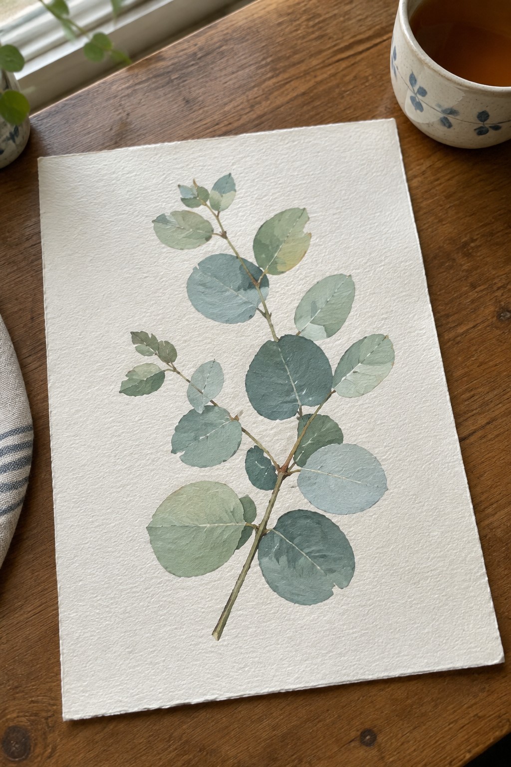 Watercolor eucalyptus branch painting on paper beside blue-patterned mug on wooden table.