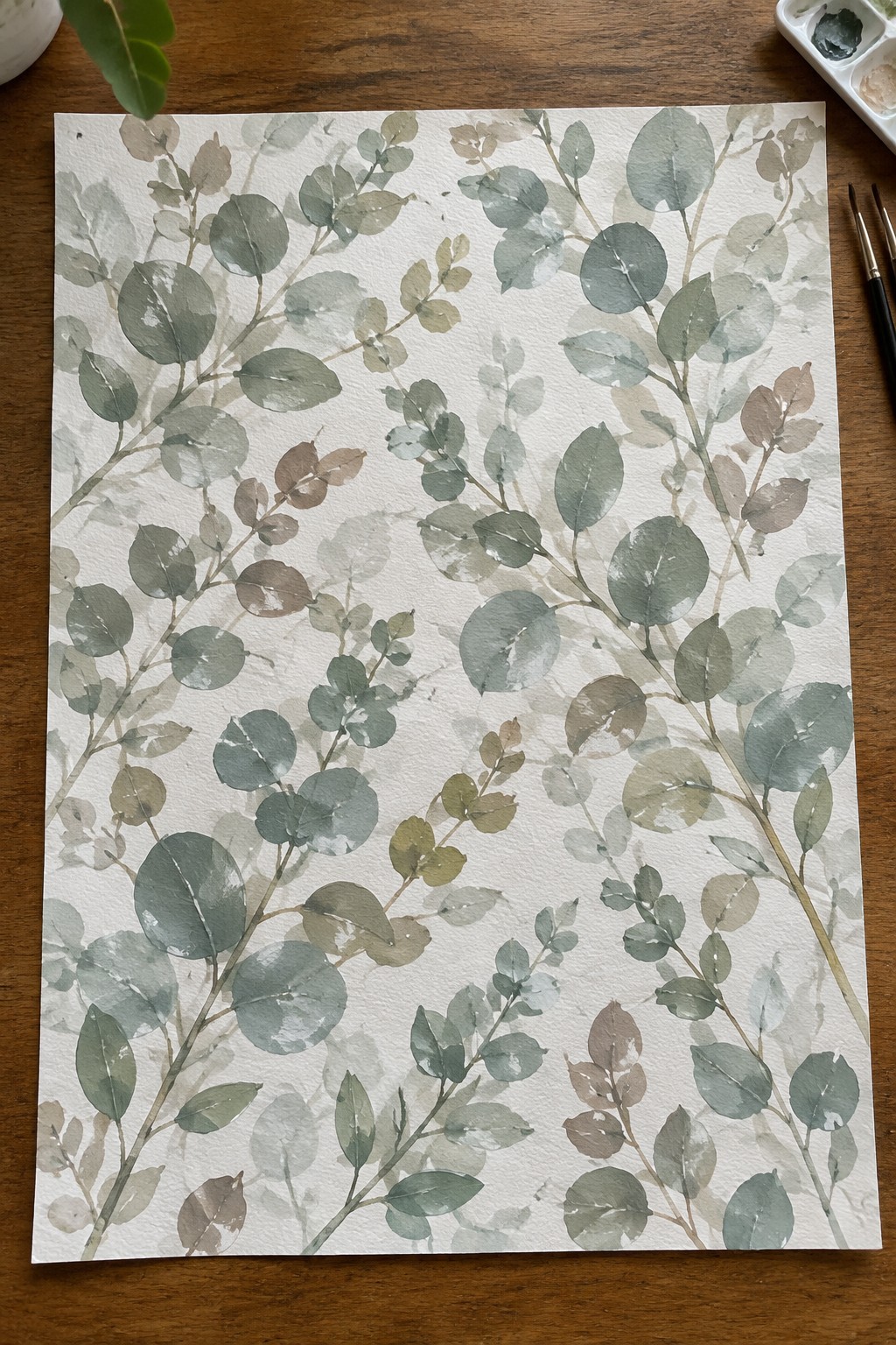 Watercolor eucalyptus branches in soft greens and browns on white paper over wood table