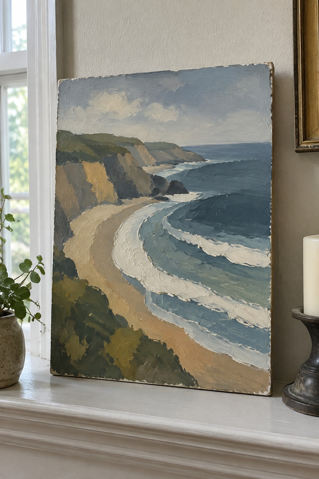 Oil painting of curving beach, blue waves, and cliffs on windowsill with plant and candle.