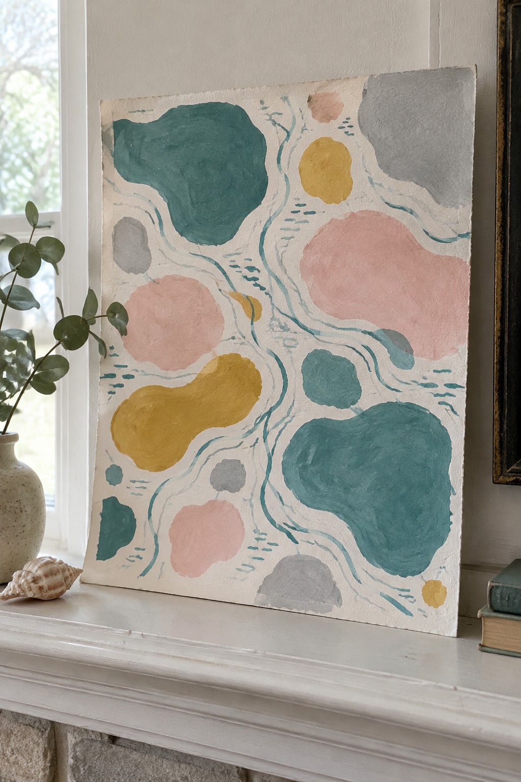 Abstract painting with teal pink yellow shapes on white mantel beside vase and books
