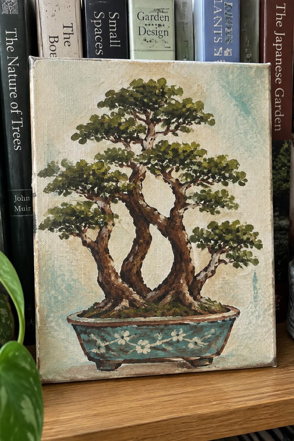A painting of a multi-trunk bonsai tree with green foliage in a teal pot decorated with white flowers, placed on a wooden surface in front of books.