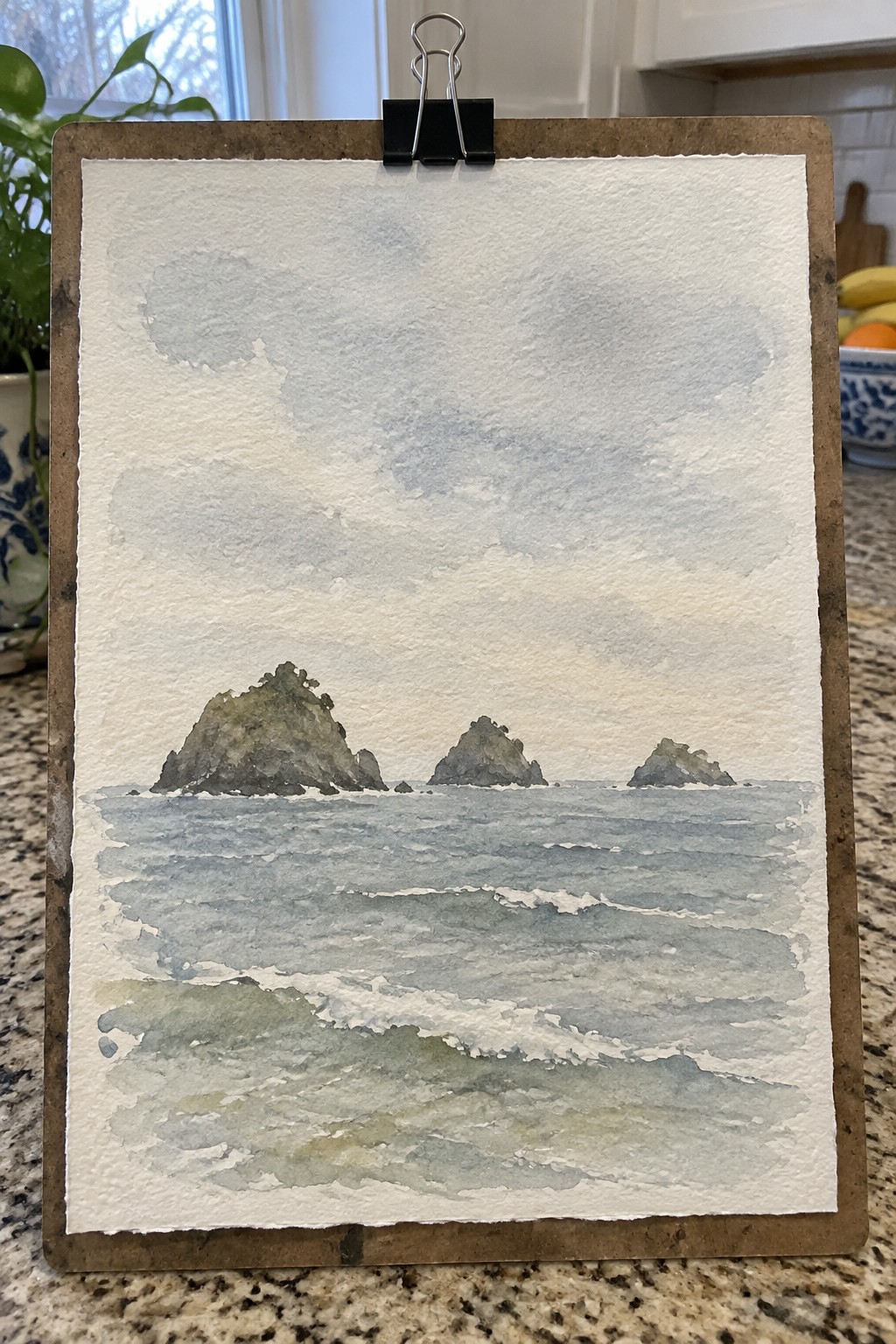 Watercolor of three rocky islands in blue sea with waves under cloudy sky on clipboard.