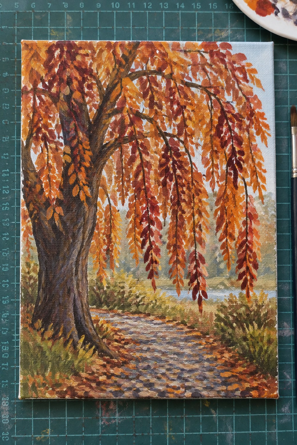 Autumn painting on canvas with orange-red tree, leaf path, brush on green mat.