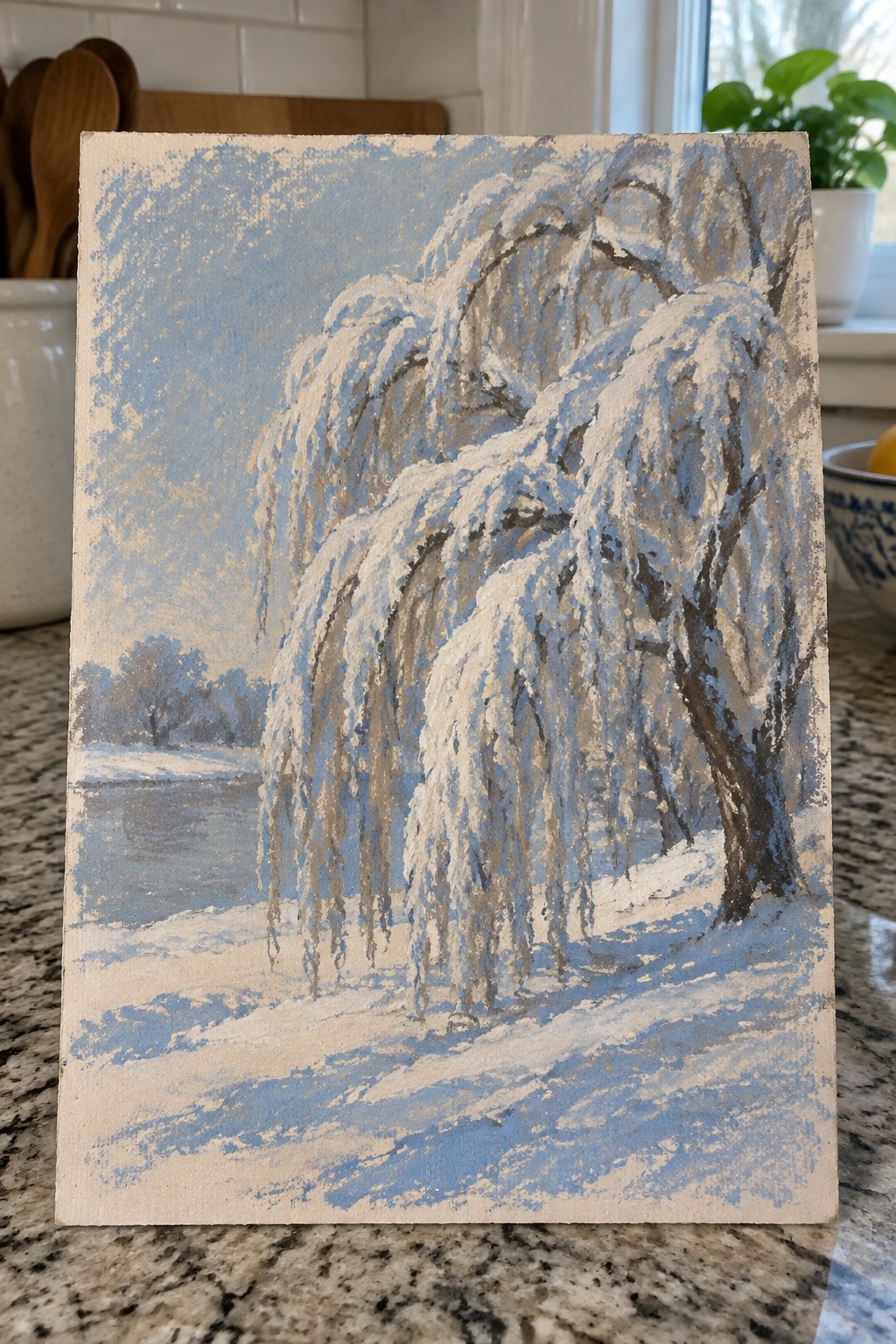 Snow-covered weeping willow painting beside frozen water on kitchen countertop