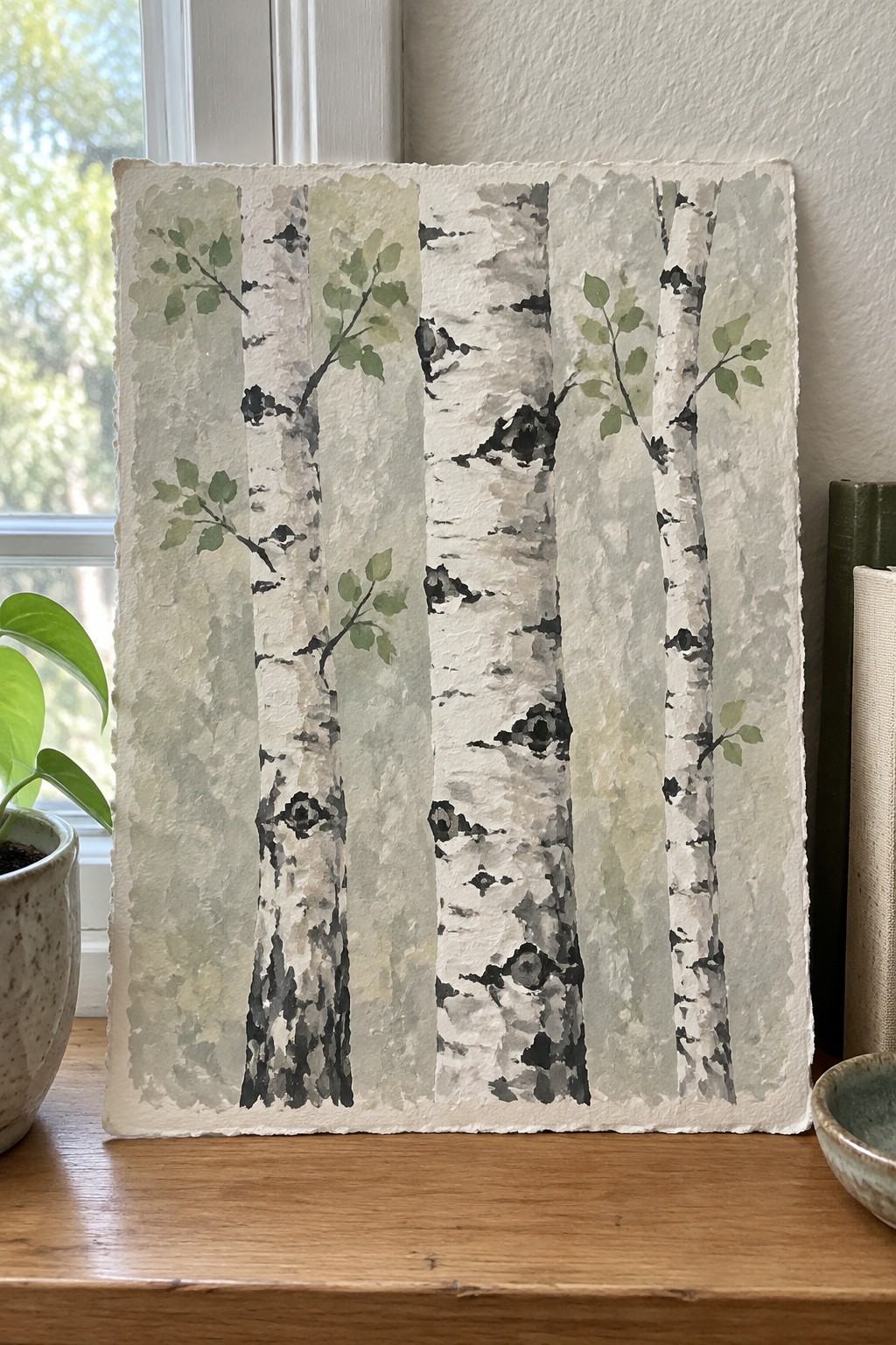 Watercolor painting of white birch trees with green leaves on textured paper by a window.