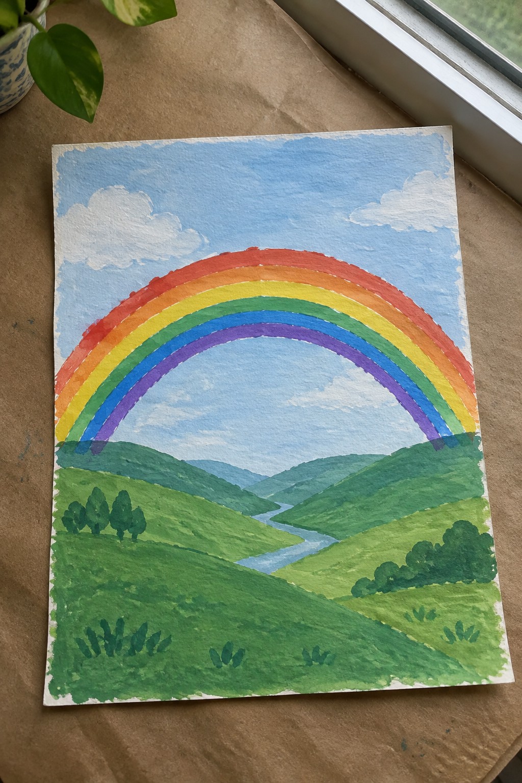 Watercolor painting of rainbow arching over green hills with winding river and blue sky.