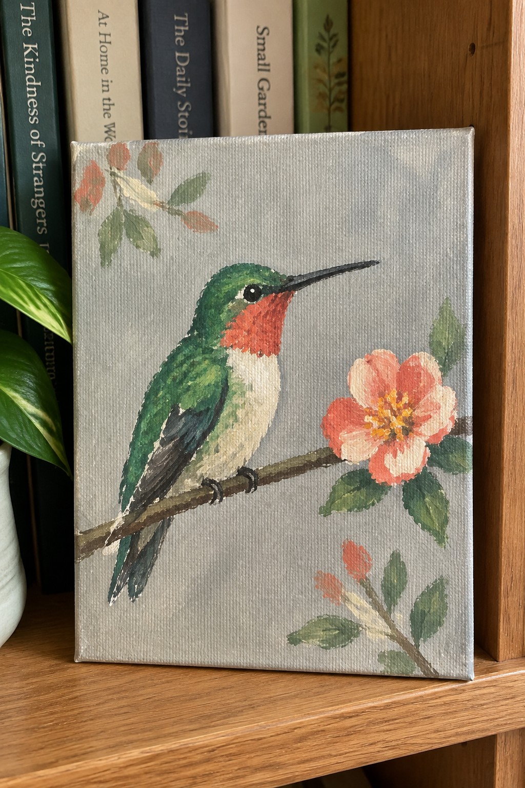 Painted canvas of green hummingbird perched on branch beside pink flower, books behind