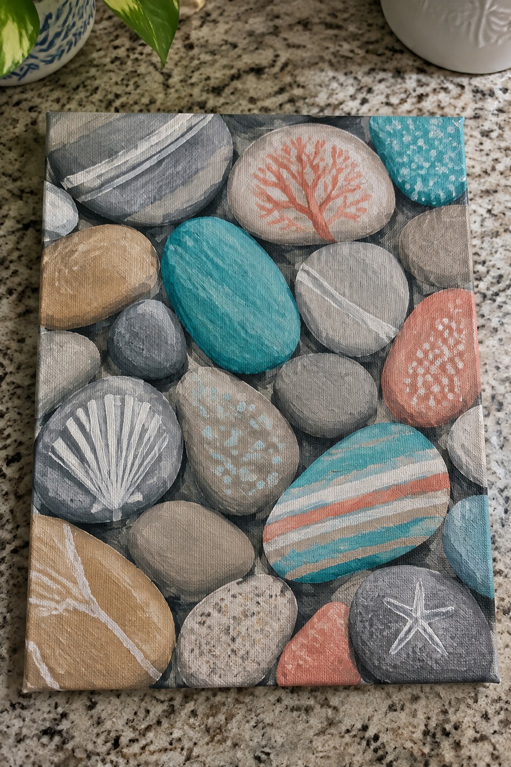 A canvas painting of many painted stones in gray, teal, brown, and coral tones, each with different patterns including stripes, dots, a shell, a starfish, and coral, arranged closely together.