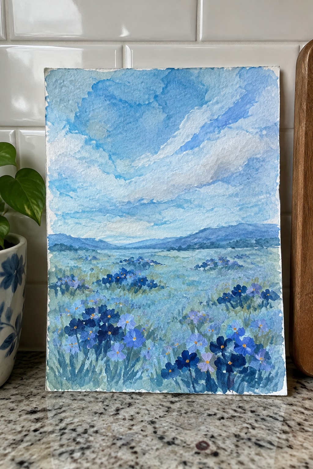 A watercolor painting of a blue flower meadow with distant hills and a cloudy sky on a kitchen counter.