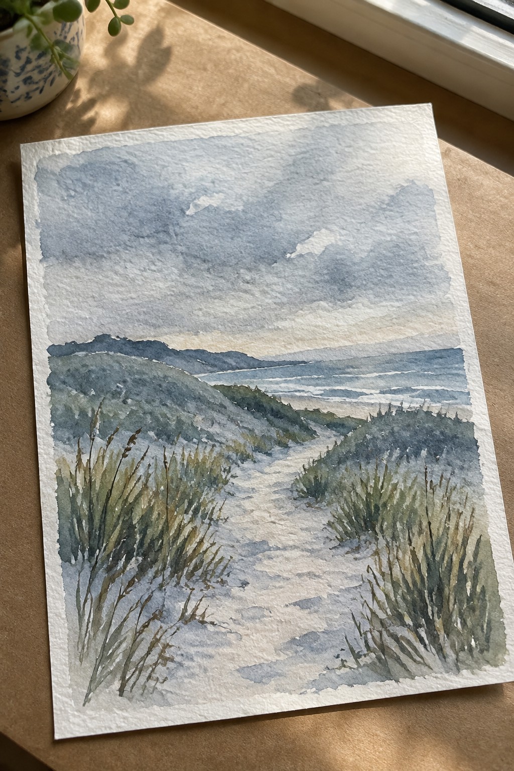 Watercolor of sandy dune path winding through grasses to ocean under cloudy sky.