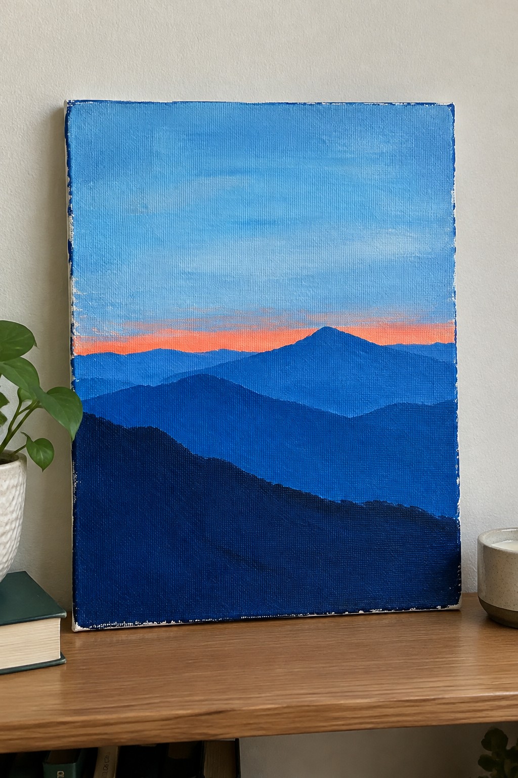 Layered blue mountains with orange sunset on wooden table beside plant and book