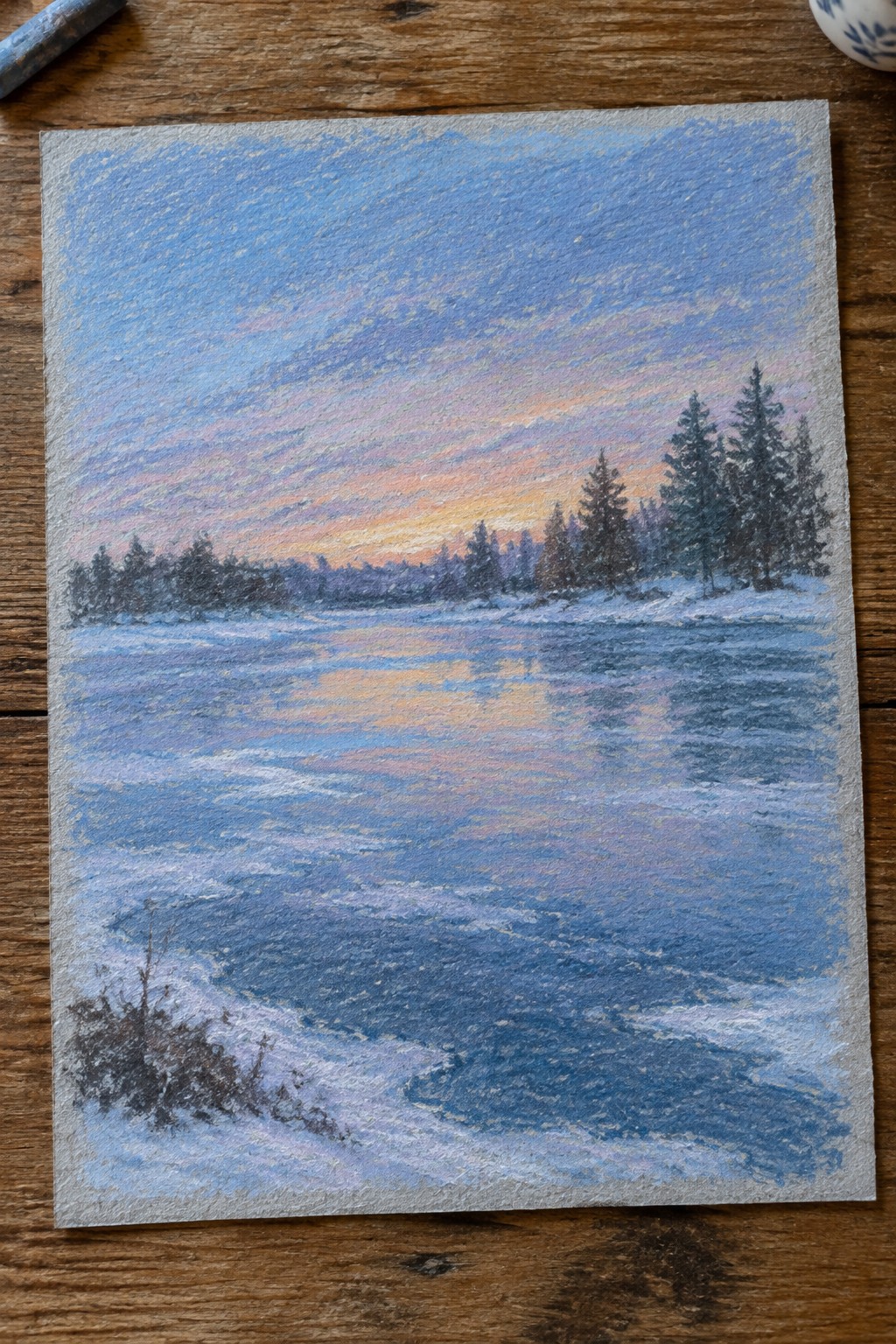 A soft pastel painting of a snowy winter landscape shows a sunset reflecting on a frozen lake with pine trees along the distant shore.